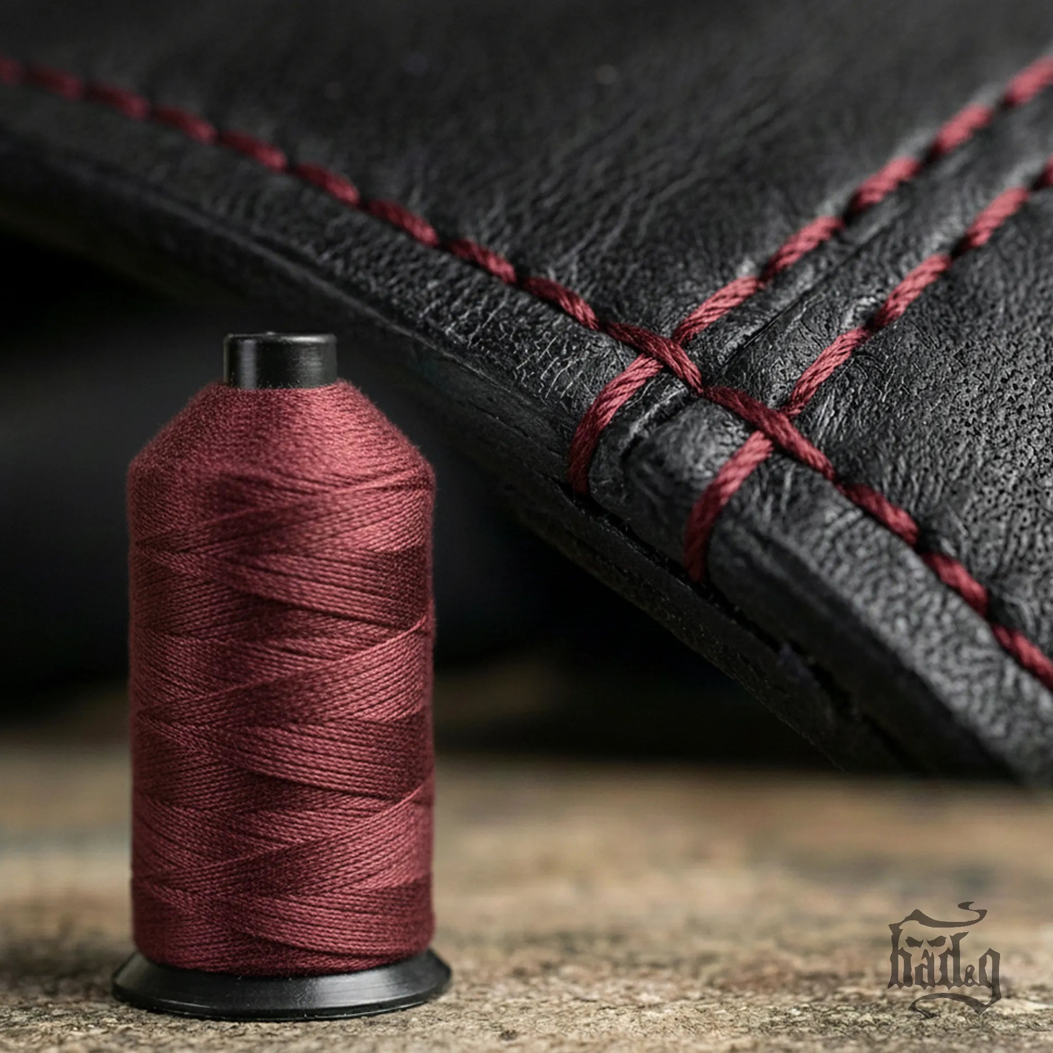Spool of burgundy thread next to a piece of black leather with burgundy stitching.